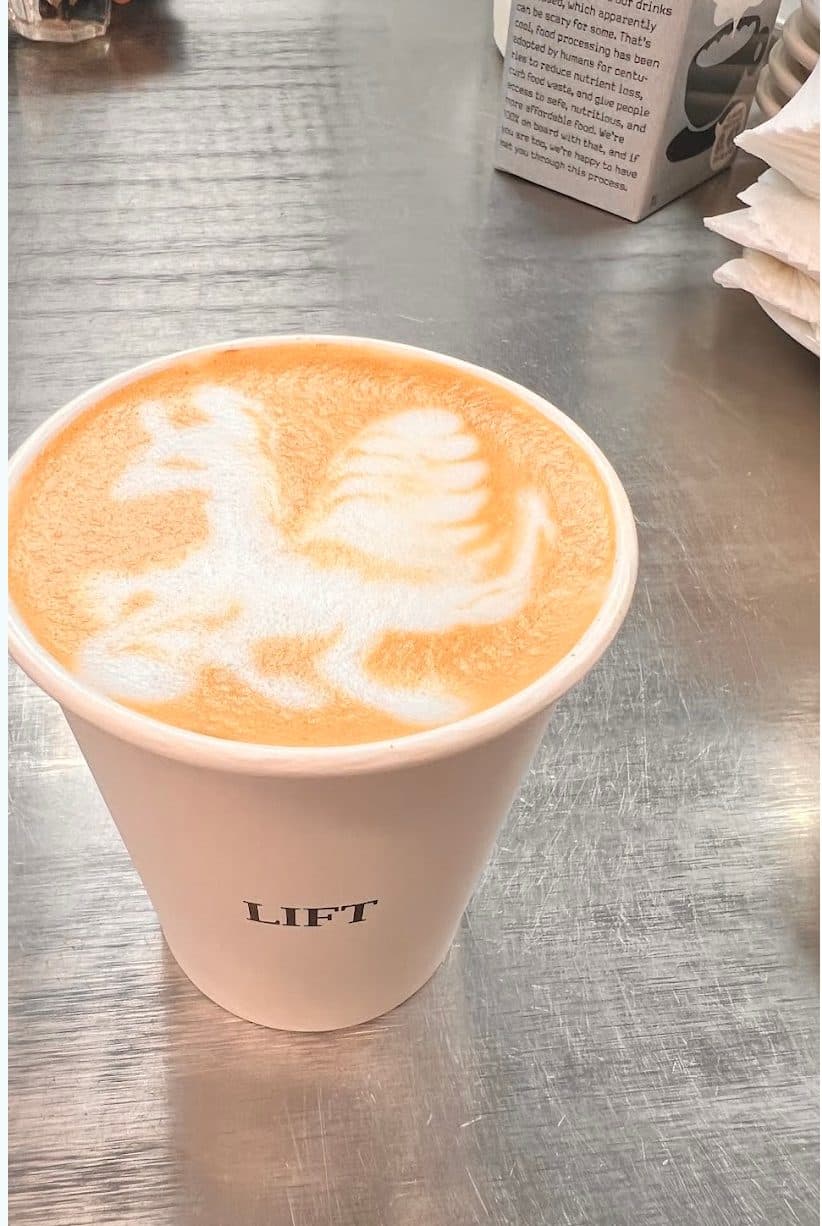 A LIFT paper cup of latte with playful latte art in the foam, on a steel counter.